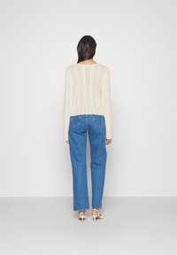 Cremfarbener Strickpullover mit einem Blättermuster, kombiniert mit blauen geraden Jeans. Weiße Absatzschuhe schaffen einen Kontrast. Sichtbare Nahtdetails.
