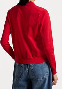 Cardigan en laine rouge tricoté avec un col rond, des poignets et un ourlet côtelés, associé à un pantalon en denim. Présente une texture lisse et une coupe ajustée.