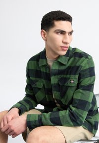 Grøn og sort ternet flannel skjorte med to frontlommer, sorte knapper og et mærke. Båret over en beige skjorte.