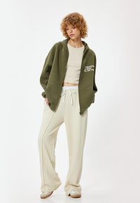 Olivengroene hoodie met ritssluiting en voorzakken, witte tekst, lichtbeige crop top en romige wijde broeken. Glad katoenen stofmix.