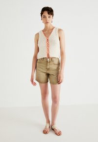 Gebreide tanktop in het wit met een roze gevlochten ontwerp, gecombineerd met beige denimshorts. Het model draagt beige gevlochten sandalen en staat tegen een effen achtergrond.