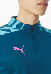 Türkises Sportshirt mit Reißverschlusskragen, der ein blaues geometrisches Muster hat, sowie einem rosa Puma-Logo auf der Brust.