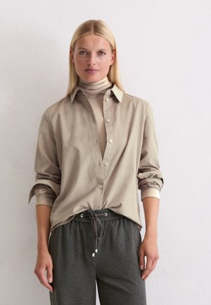 Blonde woman wearing a beige turtleneck layered with a buttoned, rolled-up sleeve shirt and dark grey drawstring pants standing against a white wall.