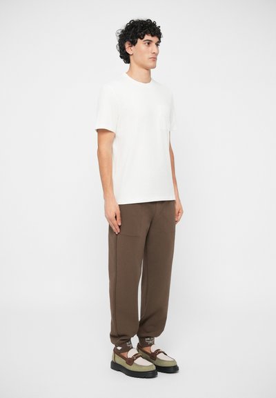 Young man wearing a white short-sleeve shirt, brown relaxed pants, patterned brown socks, and two-tone green and brown loafers.