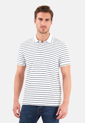 Man with short brown hair wearing a white polo shirt with thin black horizontal stripes and blue jeans, standing against a plain background.