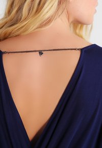 Vaalea nainen, jolla on päällä tummansininen V-taustatoppi, jossa on ohut musta ketju ja pieni kolmion muotoinen riipus yläselässä.