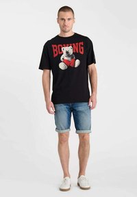 Camiseta negra con un gráfico de un oso con pantalones cortos rojos, combinada con pantalones cortos de denim claros. El modelo lleva zapatillas blancas; fondo liso.