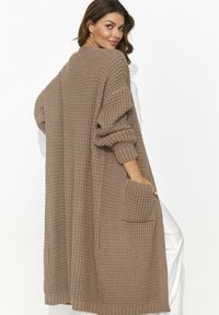 Długi, oversize sweter w miękkim brązowym dzianinie o teksturowanym wzorze, z szerokimi rękawami i przednią kieszenią. Przytulny i otwarty krój.