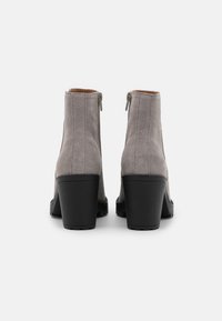 Botas de tobillo de gamuza gris con un tacón apilado negro y detalle de cremallera en la parte posterior. Textura suave con acentos de costura vertical.