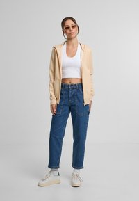 Sudadera con cremallera beige sobre un top corto blanco, combinada con unos vaqueros de tiro alto azul y zapatillas blancas con detalles verdes.