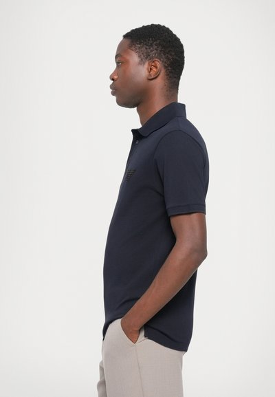 Jūras zila polo krekls, kas izgatavots no gludas auduma, ar īsu apkakli, trim pogām un teksturētu logo uz krūškurvja. Šaura piegriezuma dizains.