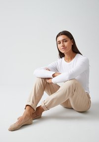 Chemise blanche à manches longues, pantalon beige retroussé, et chaussures légères. Sujet assis les bras croisés dans un cadre minimaliste.