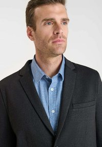Blazer nero in tessuto strutturato, con taschino sul petto e rever a punta, indossato sopra una camicia azzurro chiaro a fantasia con bottoni.