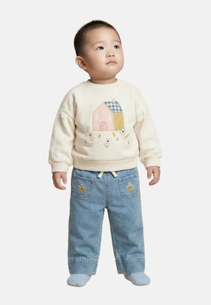 Tout-petit debout, portant un sweat-shirt crème avec un motif de maison et de fleur, un jean bleu avec une broderie de fleurs sur les poches, et des chaussettes blanches.