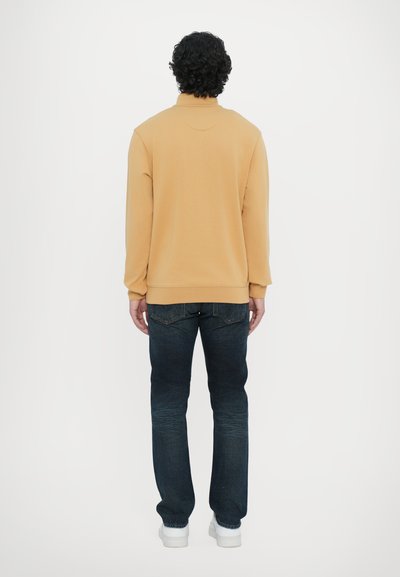 Sweatshirt beige avec un col haut, associé à un jean en denim foncé. Le sweatshirt a des poignets côtelés, et le jean a une coupe droite classique.