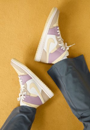 Person wearing beige, white, and lavender high-top sneakers with gray pants, feet crossed against a mustard yellow background.