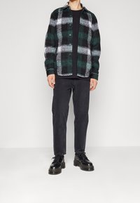 Zielono-czarna koszula flanelowa w kratę z zapięciem na guziki, noszona na czarnym T-shircie, w zestawieniu z czarnymi dżinsami i czarnymi butami.