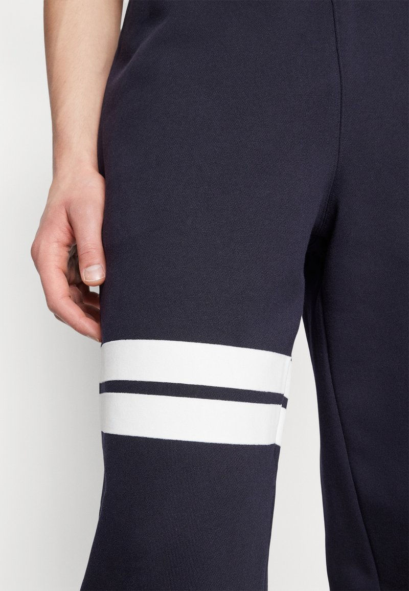 Le pantalon de survêtement bleu marine présente des rayures blanches horizontales sur la cuisse et un mélange de coton extensible pour plus de confort. Le gros plan montre le détail de la jambe.