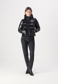 Zwarte pufferjack met een glanzende afwerking, met een capuchon, drukknoopsluitingen en elastische manchetten. Draagt het in combinatie met donkere jeans en zwarte enkellaarzen.