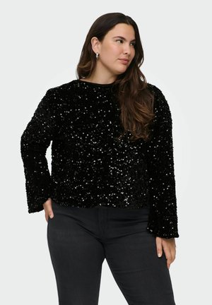 Črna bleščica z dolgimi rokavi in okroglo ovratnikom, z teksturirano površino in ohlapnim krojem. V kombinaciji s temnimi ozkimi jeans hlačami.