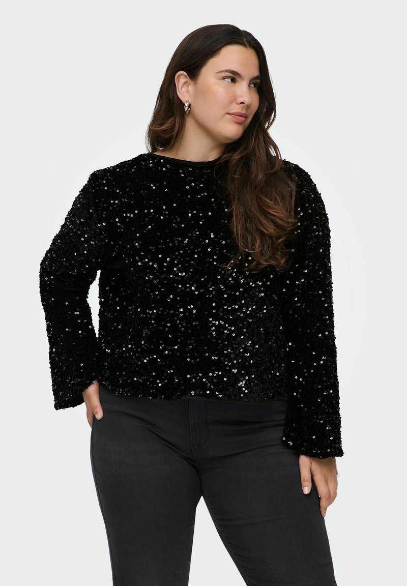 Črna bleščica z dolgimi rokavi in okroglo ovratnikom, z teksturirano površino in ohlapnim krojem. V kombinaciji s temnimi ozkimi jeans hlačami.