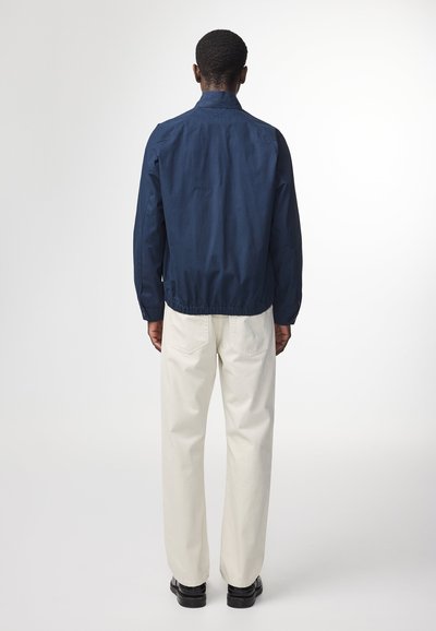 Mørkeblå jakke med krave, elastisk talje og knapmanchetter, parret med off-white bukser og sorte sko. Rent, minimalistisk design.