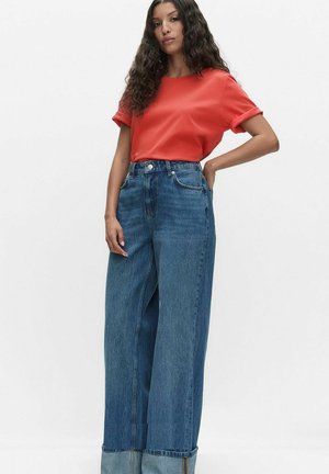 Kvinde med langt krøllet hår iført en rød kortærmet top, der er stoppet ned i højtaljede, vide blå jeans, poserer mod en hvid baggrund.