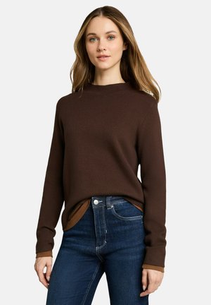 Vrouw met lichtbruin haar die een donkerbruine trui met lange mouwen draagt over een lichtere bruine blouse, die in een donkerblauwe spijkerbroek is gestopt.