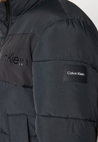 Juodas pūkinis švarkas su blizgiu audiniu, su Calvin Klein logotipu krūtinėje ir rankovėje. Uždarymas užtrauktuku ir priekinės kišenės.
