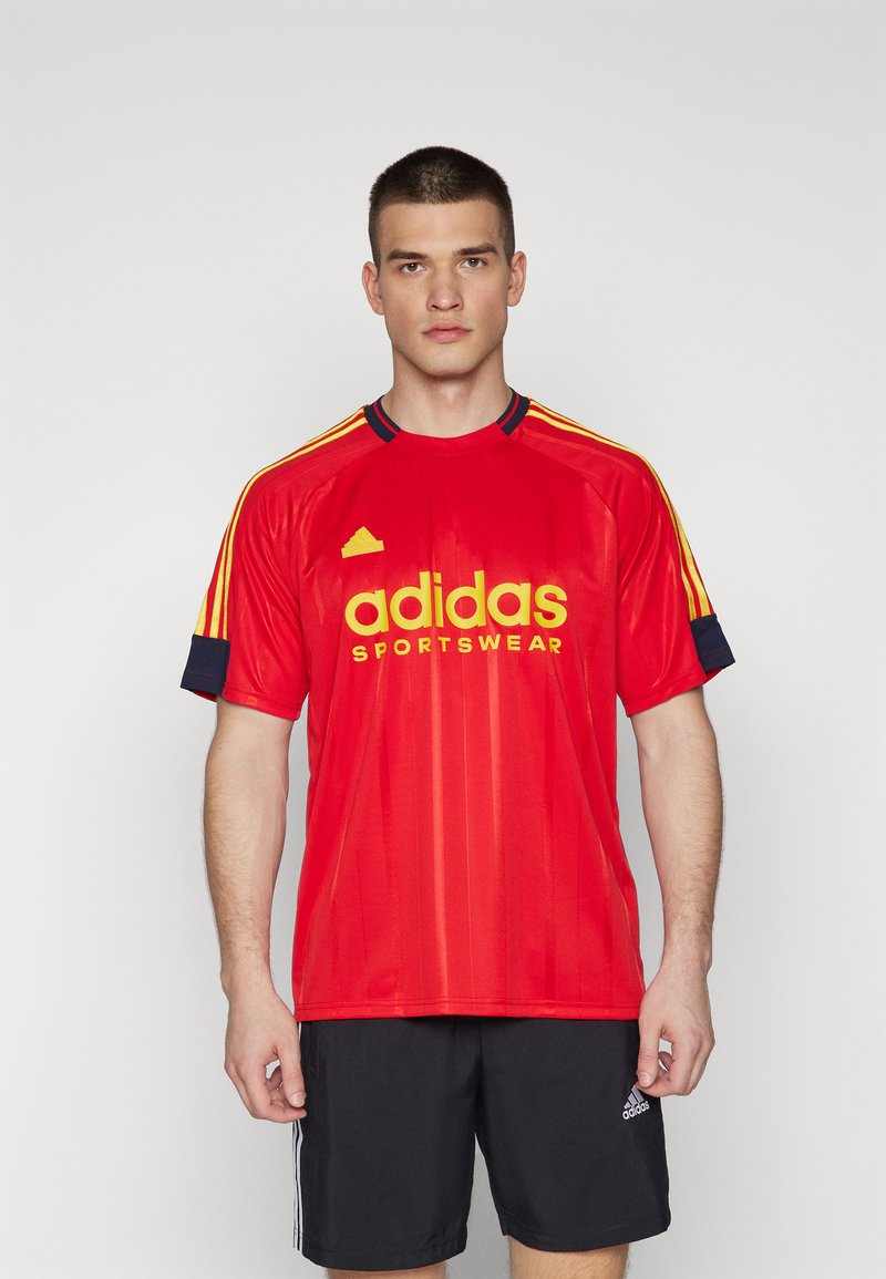 Camiseta atlética roja con el logo amarillo "adidas SPORTSWEAR". Presenta rayas horizontales en las mangas y un cuello negro. La textura de la tela es suave.