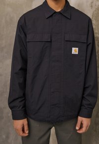 Navy blue jacket features a button-up front, two chest pockets, and a small logo patch. Made of a smooth, lightweight fabric.