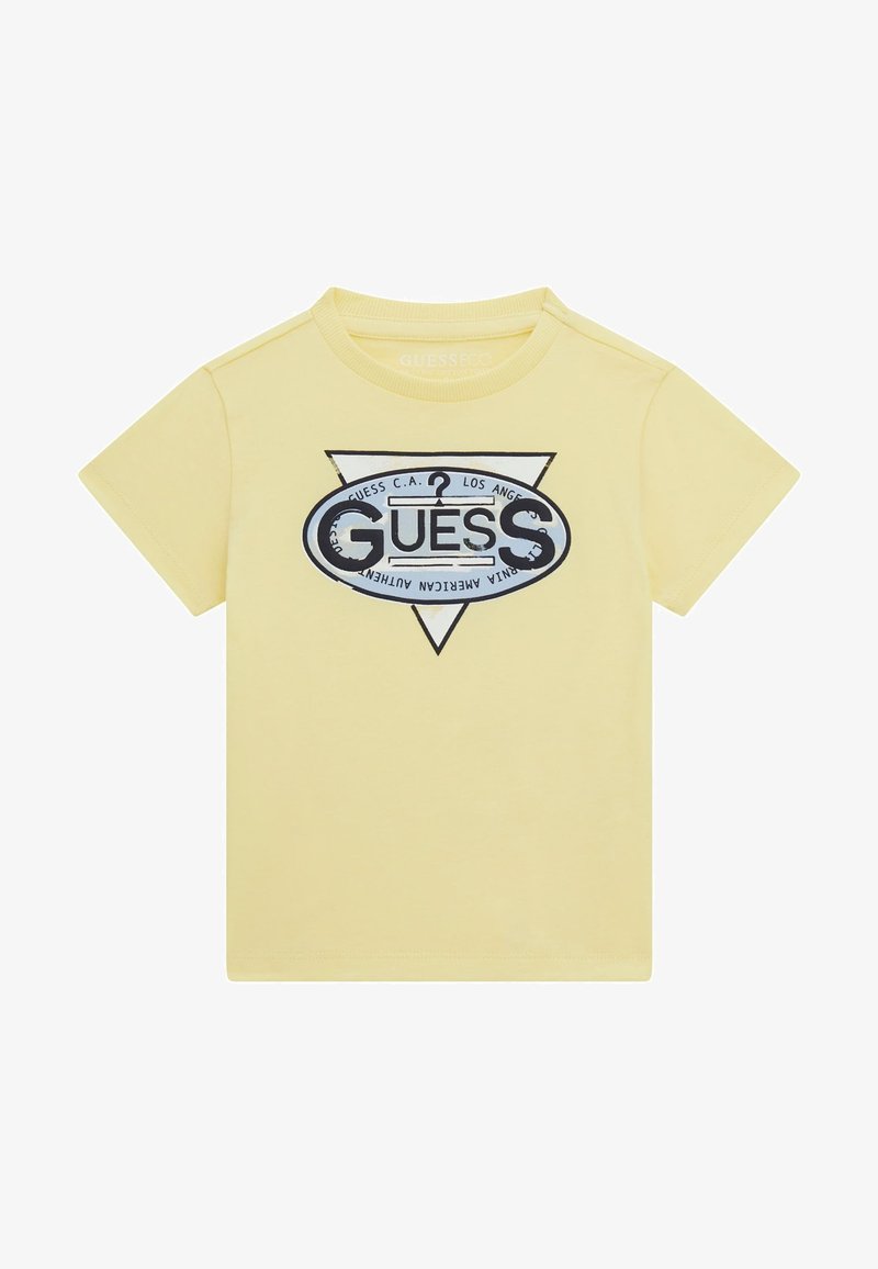 Sárga pamut póló, háromszög alakú grafikával, rajta a "GUESS" szó kék és fekete színben, további szöveggel és kerek logóval kiegészítve.