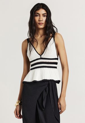 Donna con capelli lunghi e scuri, che indossa un top bianco in crochet senza maniche con righe nere e una gonna nera avvolgente, accessoriata con braccialetti d'oro.
