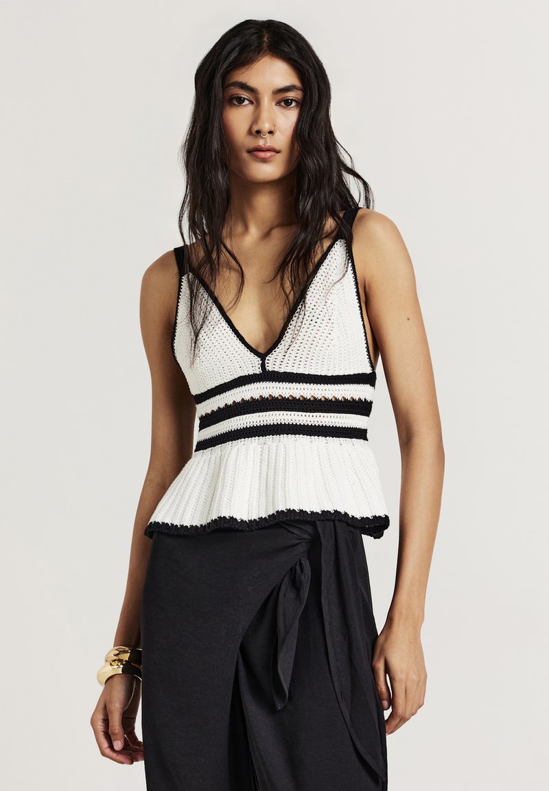 Donna con capelli lunghi e scuri, che indossa un top bianco in crochet senza maniche con righe nere e una gonna nera avvolgente, accessoriata con braccialetti d'oro.