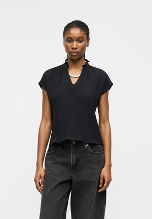 Jeune femme aux cheveux tressés porte un haut noir texturé à manches courtes et un jean sombre taille haute, debout devant un fond clair.