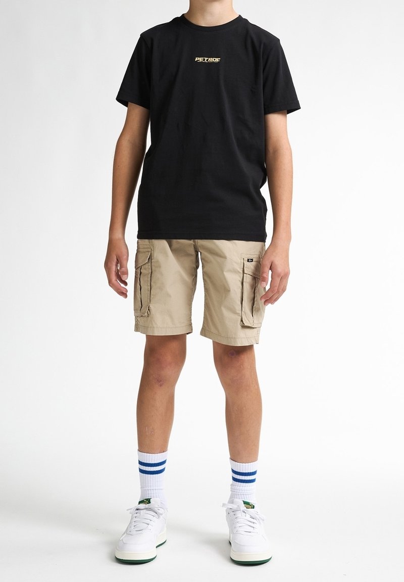 Enfant portant un t-shirt noir à manches courtes, un short cargo beige, des baskets blanches et des chaussettes blanches avec des rayures bleues, debout sur un fond blanc.