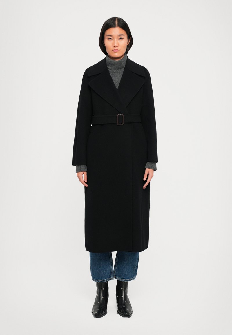 Femme portant un long manteau noir ceinturé sur un pull gris à col roulé, un jean bleu court, et des bottines noires avec œillets en métal, se tenant droite.