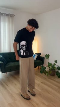 Νέος άνδρας με μπεζ παντελόνι και μαύρο γραφιστικό t-shirt, στέκεται σε σαλονάκι με πράσινο καναπέ, φυτά και ξύλινο πάτωμα.