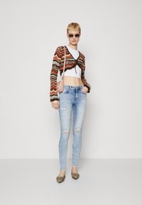 Knit cardigan with multicolored zigzag pattern, white crop top, distressed light blue skinny jeans, and metallic flat shoes. Pink handbag.