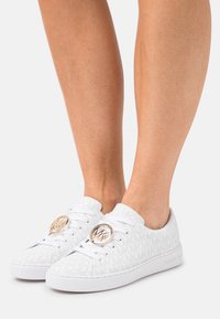 Zapatillas blancas de tela texturizada con un patrón de logo en relieve. Punta redonda, suela blanca plana y acentos circulares en tono dorado en los cordones.