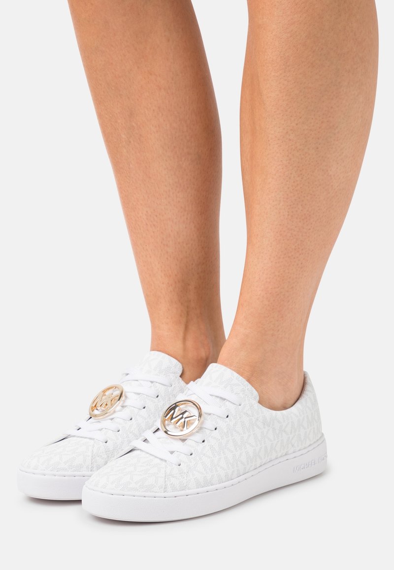 Zapatillas blancas de tela texturizada con un patrón de logo en relieve. Punta redonda, suela blanca plana y acentos circulares en tono dorado en los cordones.