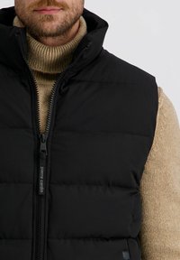 Schwarze gepolsterte Weste mit hohem Kragen und Frontreißverschluss, versehen mit einem Logo am Reißverschlusszipper. Getragen über einem hellbraunen Strickpullover.