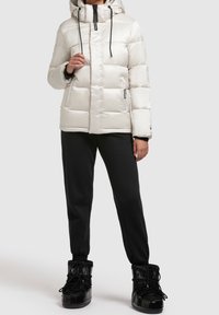 Chaqueta blanca acolchada con capucha, cordones negros, cierre delantero con cremallera, dos bolsillos laterales, combinada con pantalones deportivos negros y botas negras brillantes.