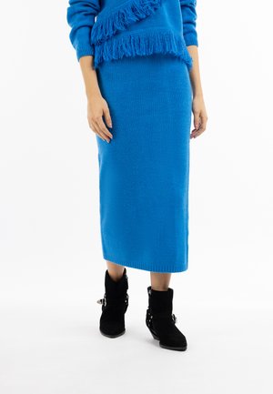 Kvinde iført en blå strikket midi-nederdel og matchende sweater med frynser og sorte ankelstøvler, stående på en hvid baggrund.