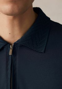 Mörkblå zip-up plagg med en texturerad krage som har ett zickzackmönster. Tyget är slätt och dragkedjan är metallisk.