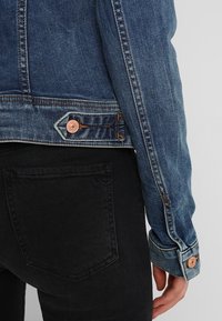 Denim jacket with a dark wash, featuring copper hardware, contrasting stitching, and a fitted design. Paired with black jeans, showing pocket detail.