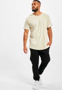 Beige T-shirt met ronde hals en korte mouwen, gecombineerd met zwarte cargobroek en witte sneakers. De outfit heeft een relaxed fit en een gladde textuur.
