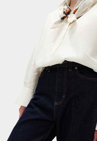 Camicia bianca a maniche lunghe con bottoni abbinata a jeans di denim blu scuro. L'outfit è completato da una sciarpa di seta a fantasia legata al collo.