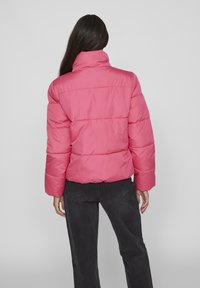 Jacket roz pal cu puf, având un guler înalt, un design matlasat și manșete cu nervuri. Purtați cu pantaloni gri închis, văzuți din spate.
