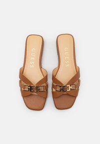 Sandales à glissière marron avec un design à lanière croisée, quincaillerie dorée et semelle plate. Texture lisse avec une semelle intérieure marquée.